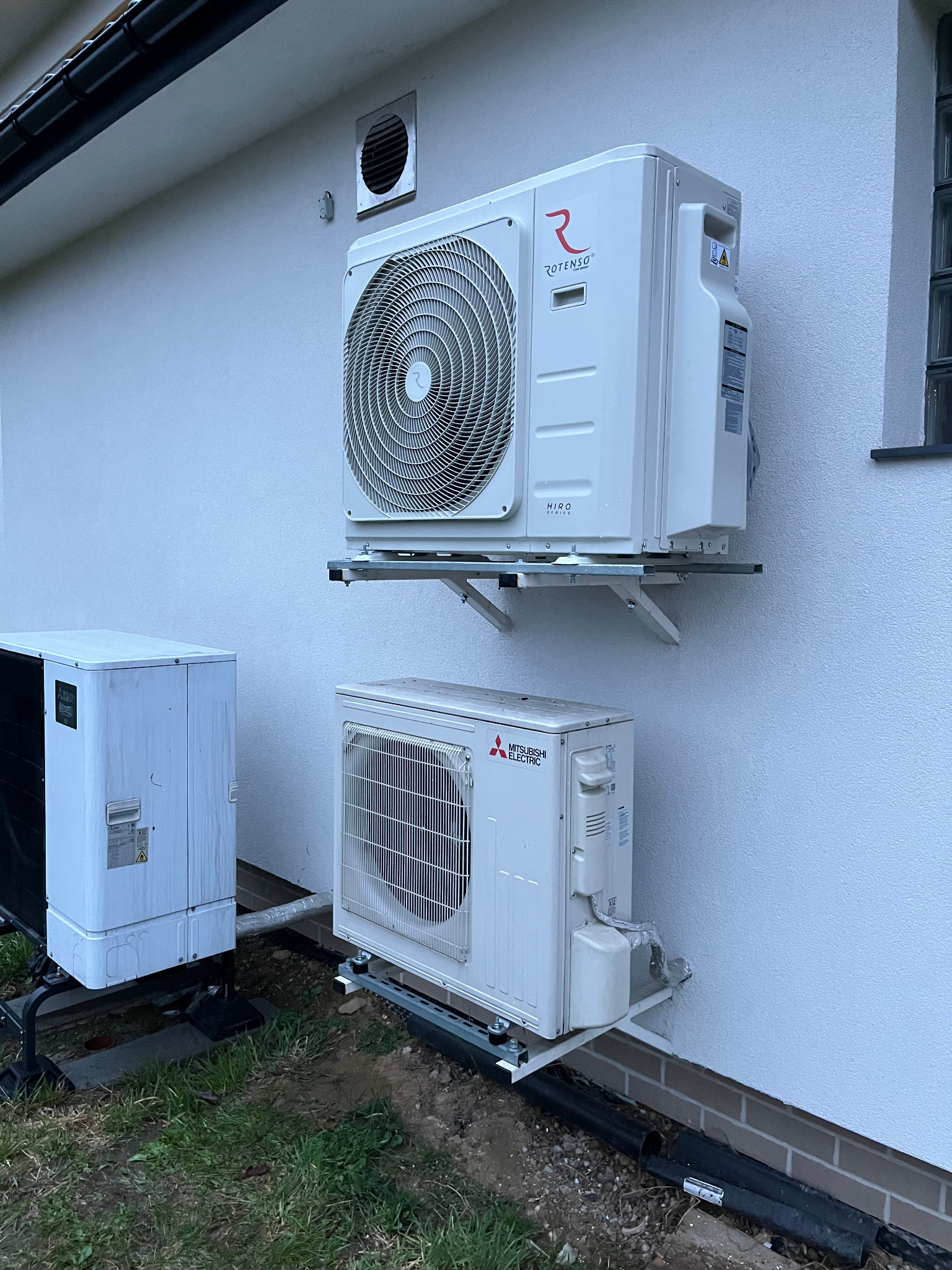 Dwa klimatyzatory Rotenso i Mitsubishi zamontowane na elewacji budynku, widoczne przewody i elementy instalacji. Zewnętrzna jednostka klimatyzacji.
