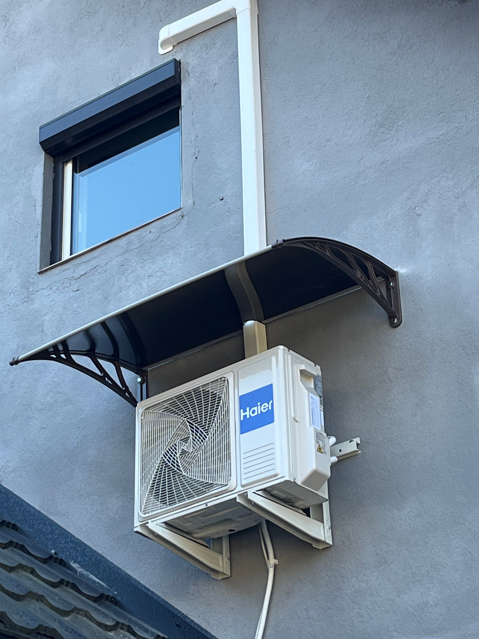 Zamontowana jednostka zewnętrzna klimatyzacji Haier na szarej elewacji budynku, osłonięta daszkiem. Widoczny fragment okna z roletą i rura spustowa.