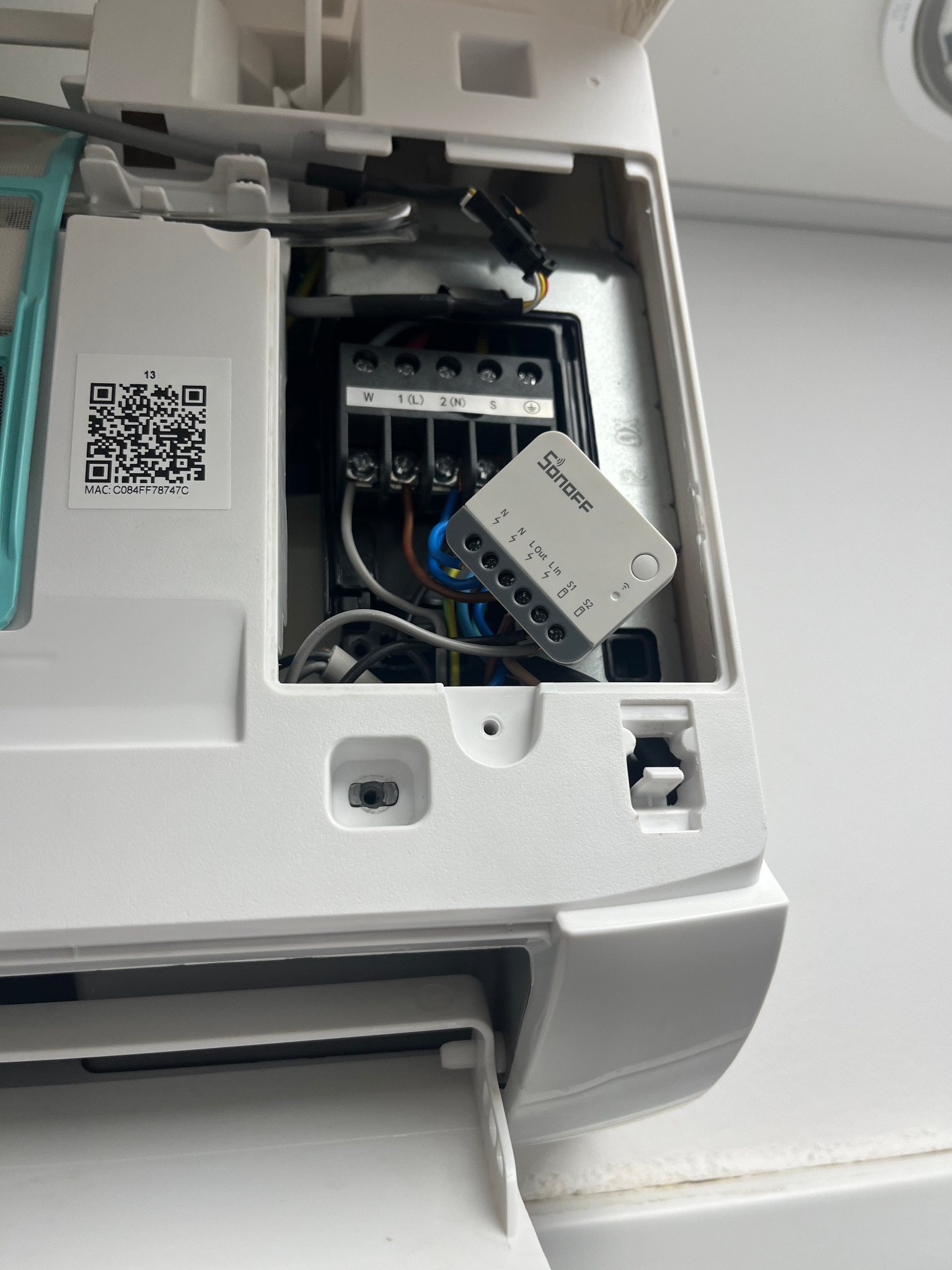 Wnętrze klimatyzatora z podłączonym modułem Sonoff. Widoczne przewody, złącza i etykieta z kodem QR. Biała obudowa urządzenia na jasnym tle.