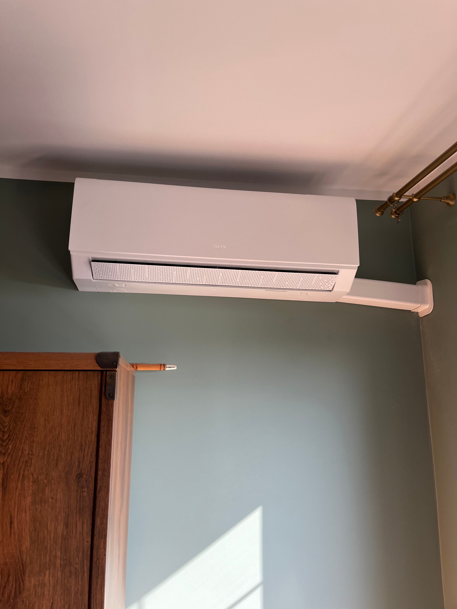 Biała klimatyzacja AUX zamontowana pod sufitem na tle ściany w odcieniach zieleni i brązu, widoczny fragment szafy i karnisza.
