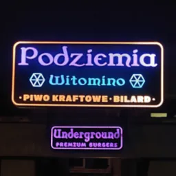 Podświetlany szyld reklamowy z napisem 'Podziemia Witomino', 'Piwo Kraftowe Bilard' oraz mniejszy szyld 'Underground Premium Burgers' w ciemnym otoczeniu.