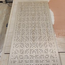 JJ CNC - Płyta z jasnego drewna z precyzyjnie wywierconymi otworami o regularnym wzorze, leżąca na stole roboczym z widocznymi śladami obróbki.