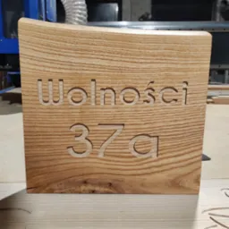 Drewniana tabliczka adresowa z wyfrezowanym napisem 'Wolności 37a', wykonana z jasnego drewna o wyraźnym usłojeniu, obróbka CNC w tle.