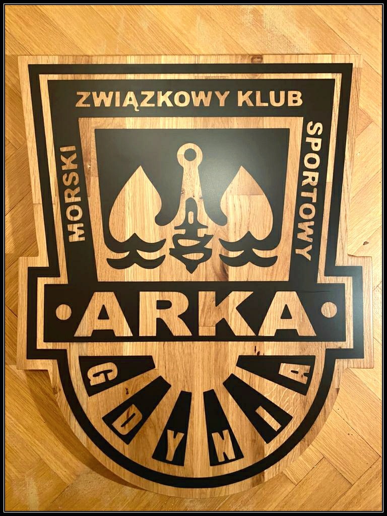 Drewniany szyld z wygrawerowanym logo 'Związkowy Klub Sportowy ARKA Morski', z kotwicą i stylizowanymi falami, leżący na drewnianej podłodze.