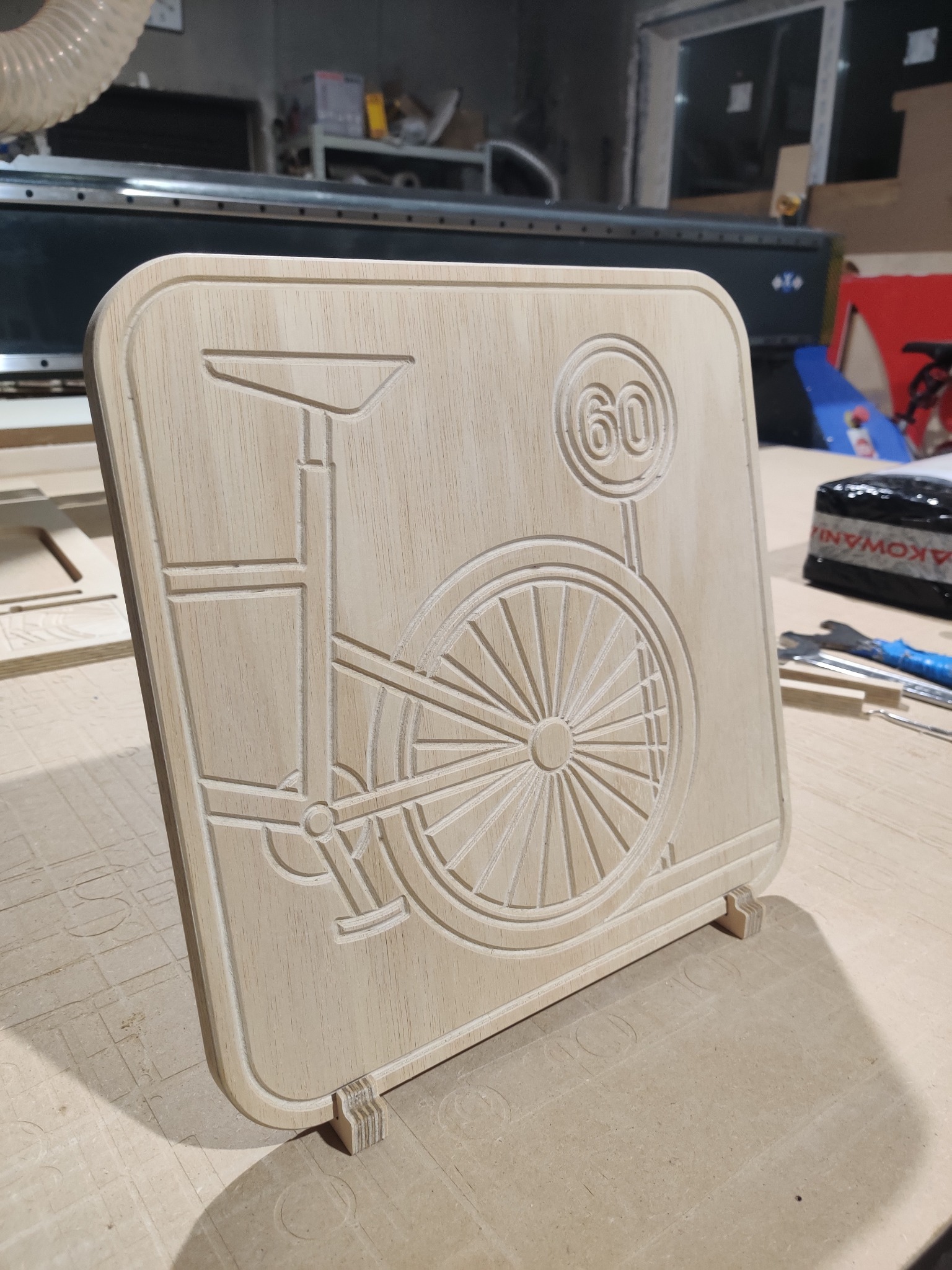Drewniana tabliczka z wygrawerowanym motywem roweru typu 'bicykl' i znakiem drogowym '60', wykonana na maszynie CNC, widoczna na blacie roboczym z widoczną maszyną CNC w tle.