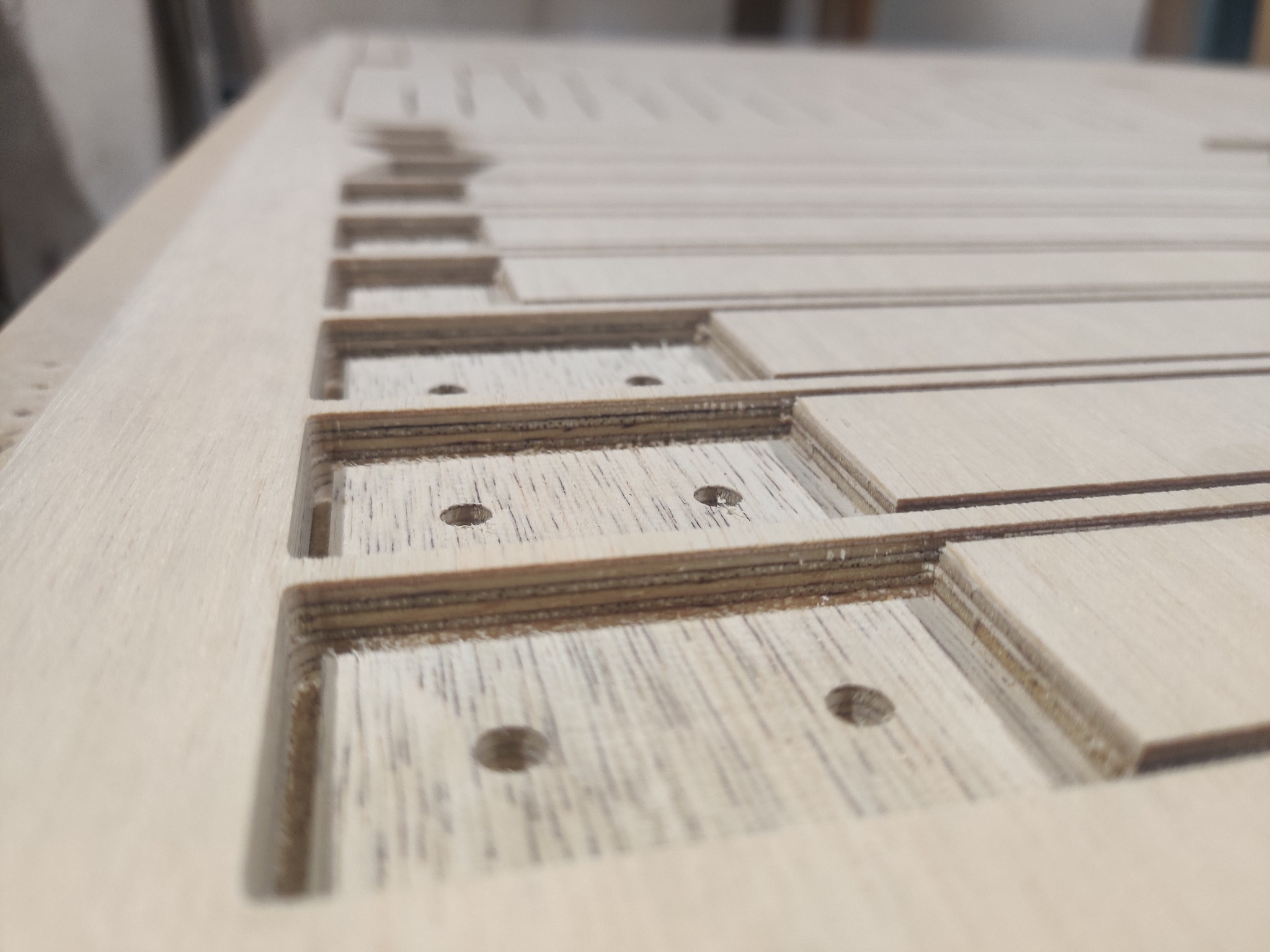 Płyta z jasnego drewna z precyzyjnie wyfrezowanymi prostokątnymi otworami i nawierconymi otworami, widoczne warstwy sklejki na krawędziach, wykonana obróbką CNC.