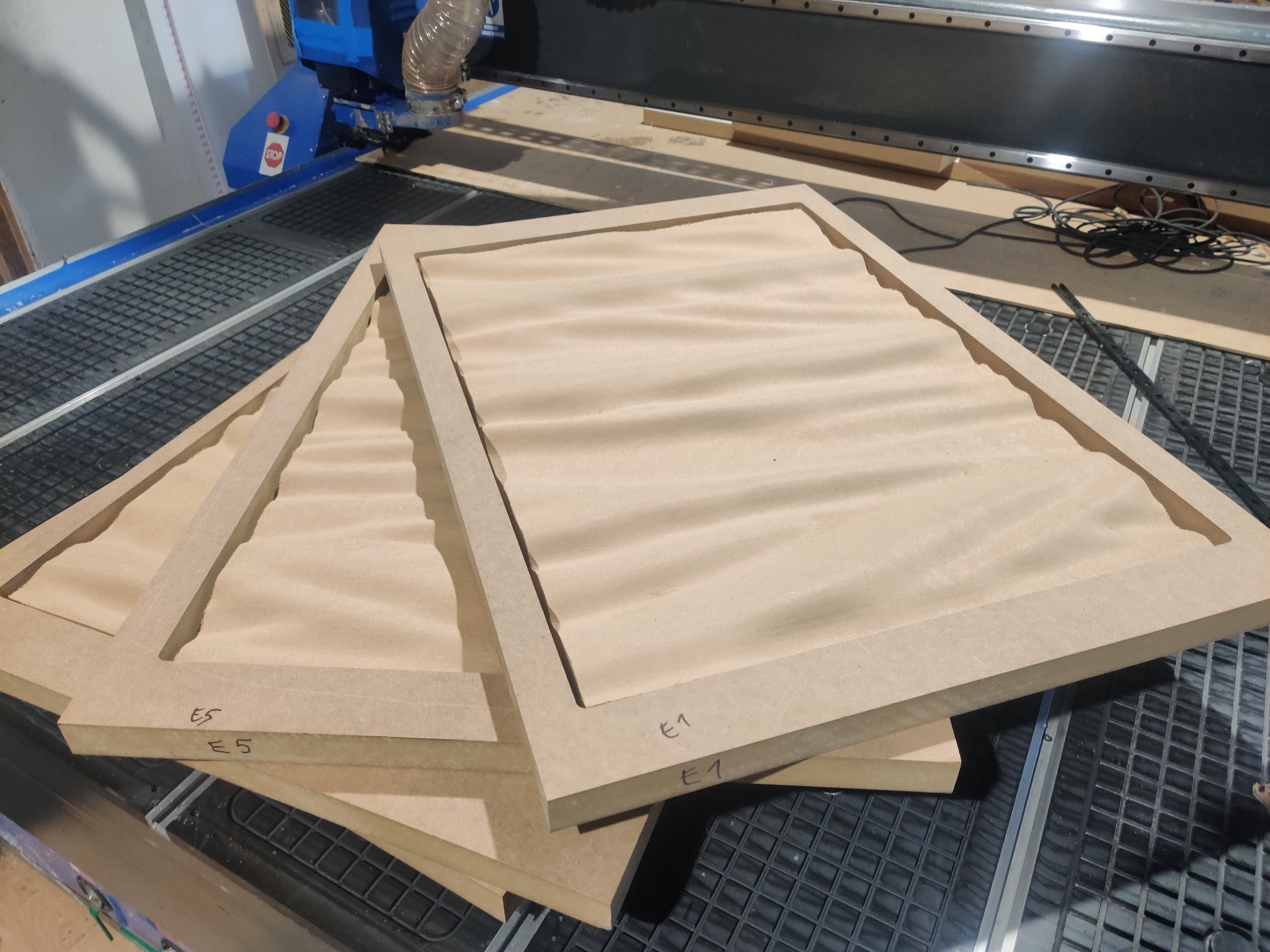 Trzy drewniane ramy z wyfrezowanymi wzorami leżące na stole frezarki CNC, oznaczone numerami E1 i E5.