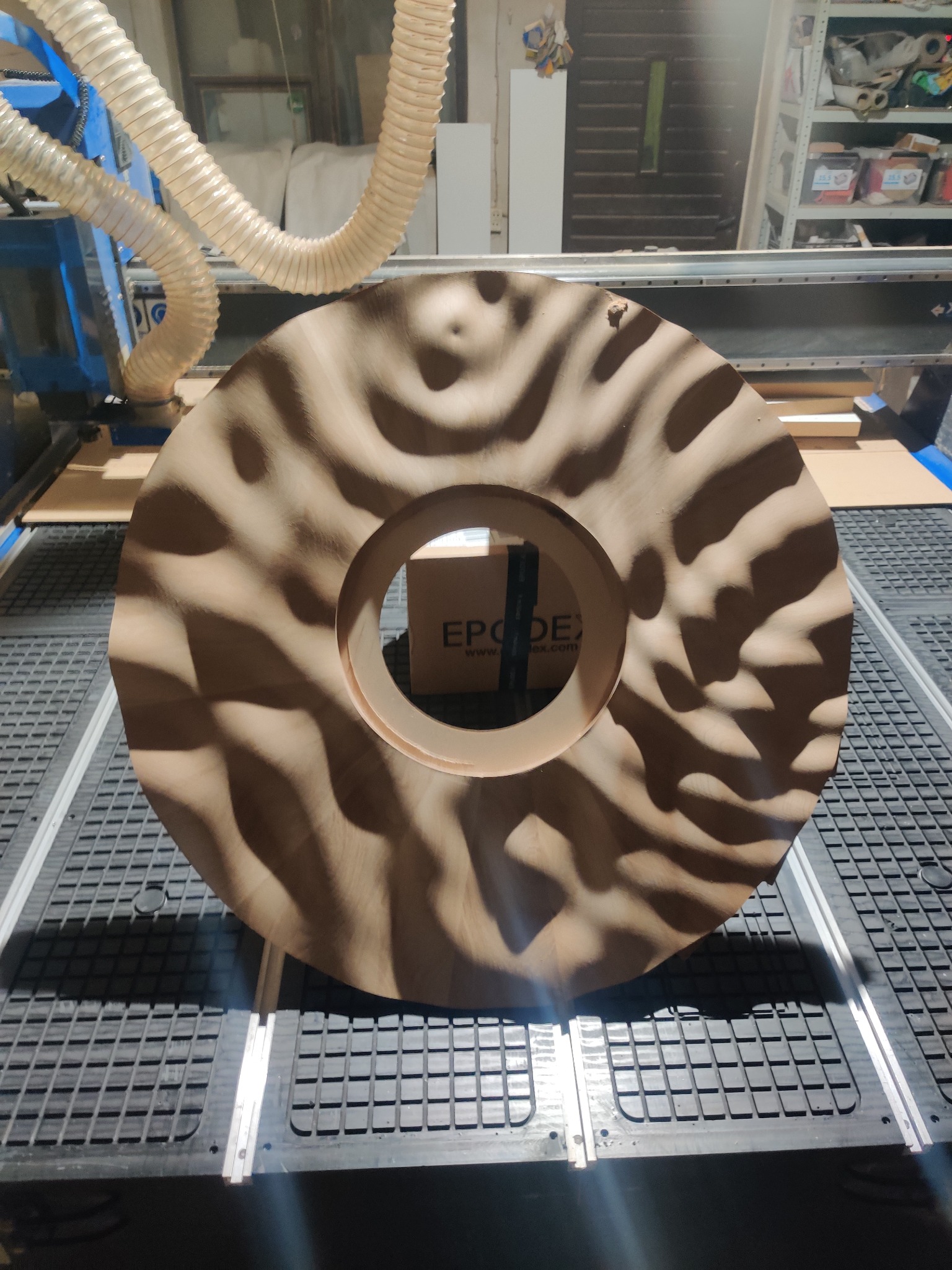 Drewniany element dekoracyjny w kształcie okręgu z nieregularnym wzorem falistym, wykonany przy użyciu frezarki CNC, leżący na stole roboczym maszyny.