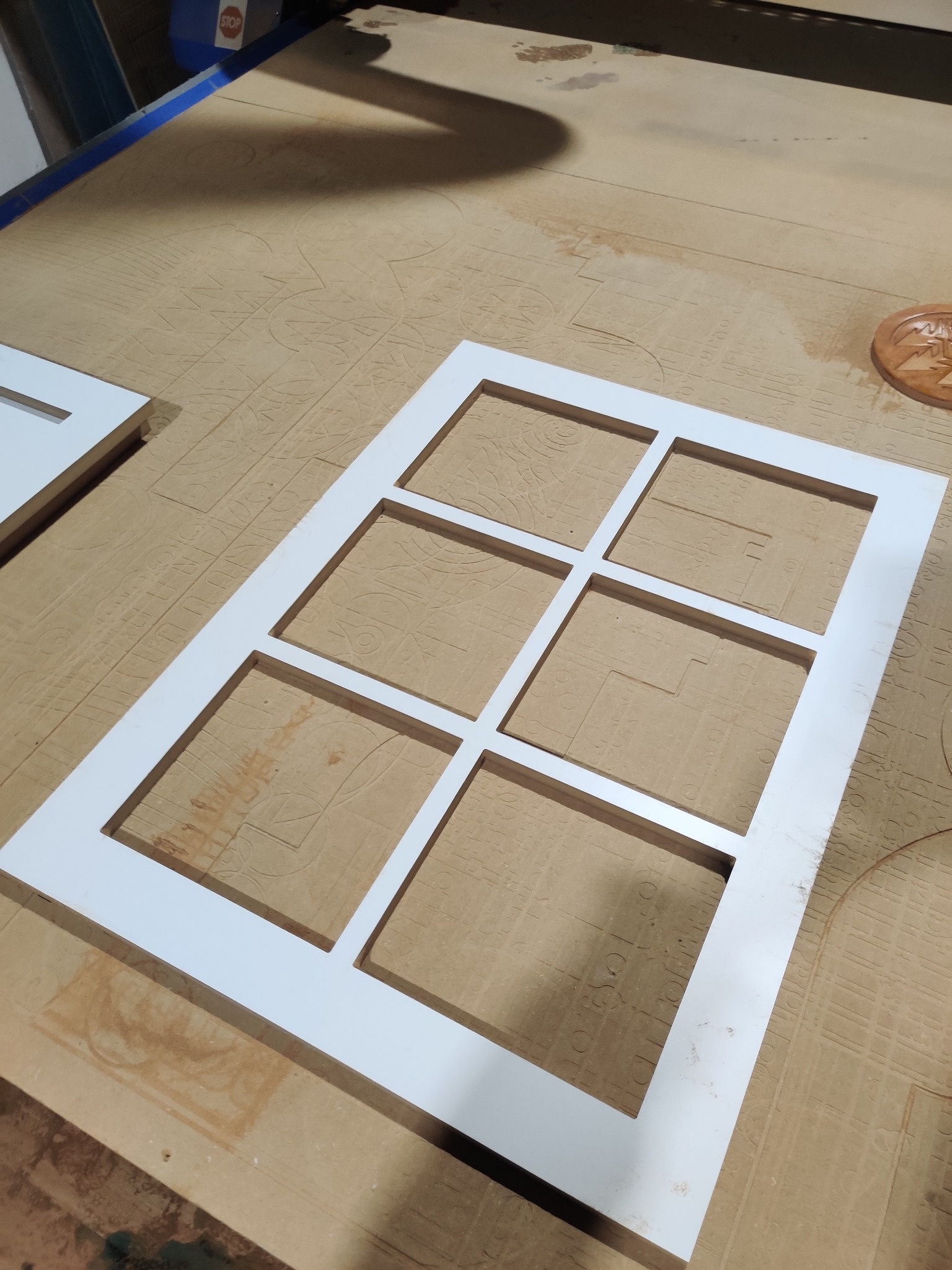 Biała rama okienna z sześcioma prostokątnymi otworami, wycięta na obrabiarce CNC, leżąca na stole roboczym z widocznymi śladami frezowania i elementami mocującymi.