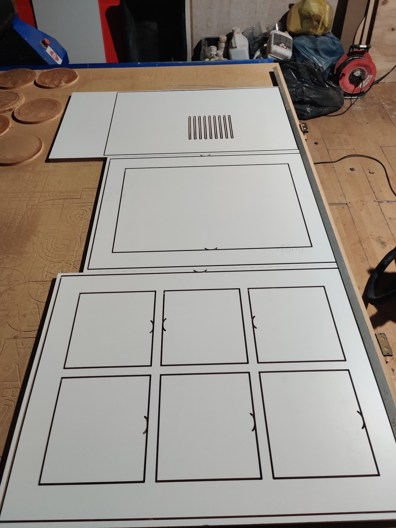 Białe, laminowane płyty z wyfrezowanymi wzorami i otworami, prawdopodobnie elementy frontów meblowych, leżące na stole warsztatowym.