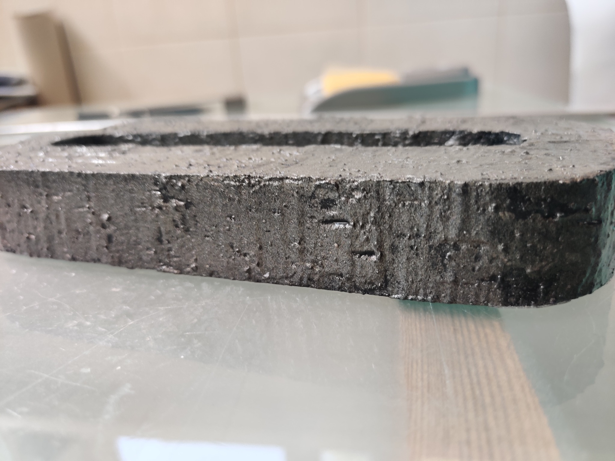 Czarny, prostokątny element z wyciętym otworem, wykonany prawdopodobnie z tworzywa sztucznego, leżący na błyszczącej powierzchni. Widoczne ślady obróbki i porowata struktura.