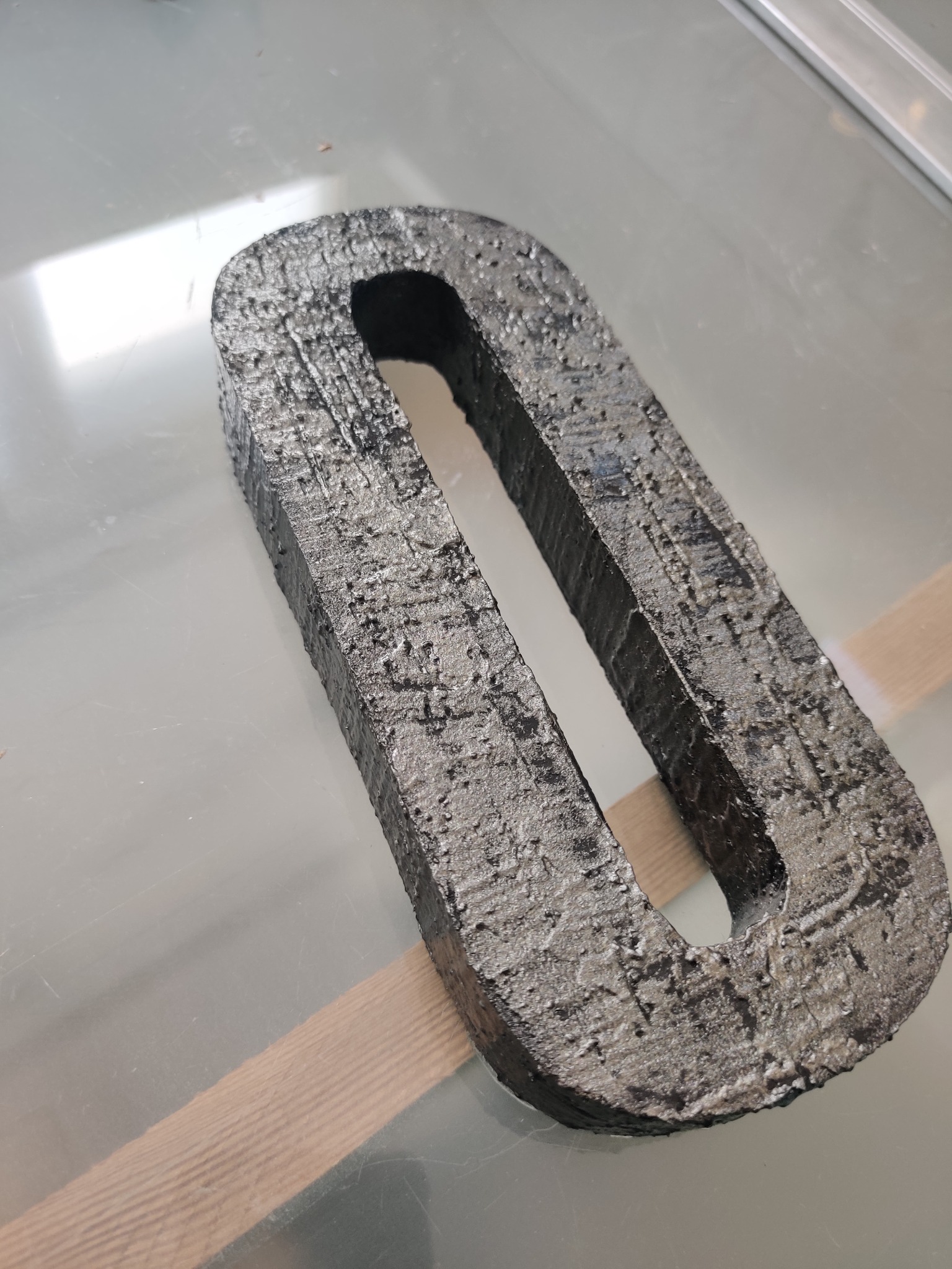 Metalowy element w kształcie litery 'O' z wyraźną, chropowatą strukturą powierzchni, leżący na szklanym blacie.