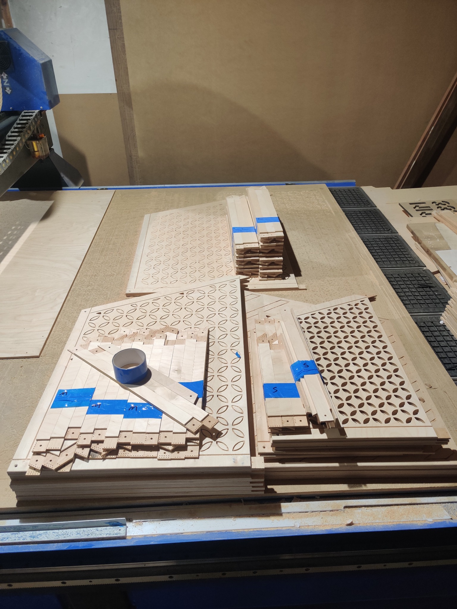 Drewniane panele dekoracyjne z wyciętymi wzorami, zabezpieczone niebieską taśmą, ułożone na stole roboczym maszyny CNC.