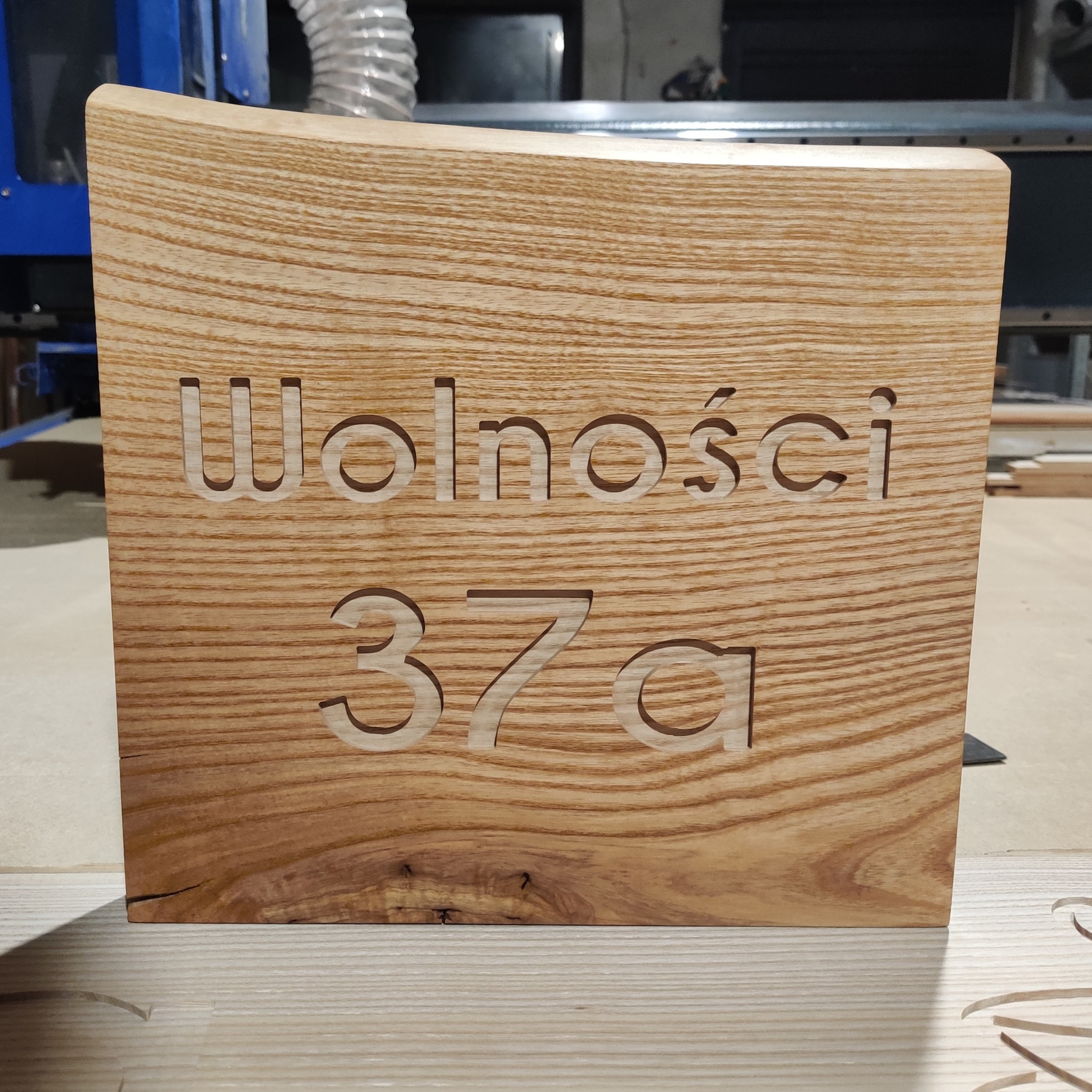 Drewniana tabliczka adresowa z wyfrezowanym napisem 'Wolności 37a', wykonana z jasnego drewna o wyraźnym usłojeniu, obróbka CNC w tle.