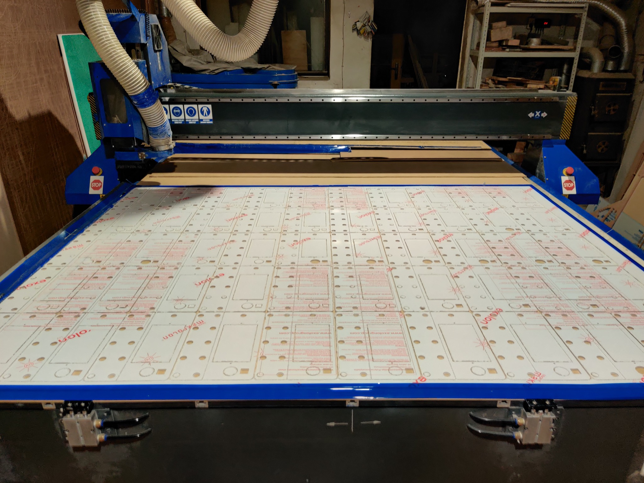 Duża frezarka CNC z białym arkuszem tworzywa sztucznego, na którym widoczne są powtarzalne wzory do wycięcia, z czerwonymi napisami producenta.