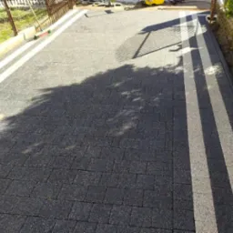 Ukończona nawierzchnia z kostki brukowej w odcieniach szarości, z jasnymi pasami. Widoczne cienie drzew i sprzęt budowlany w tle. Detal realizacji.