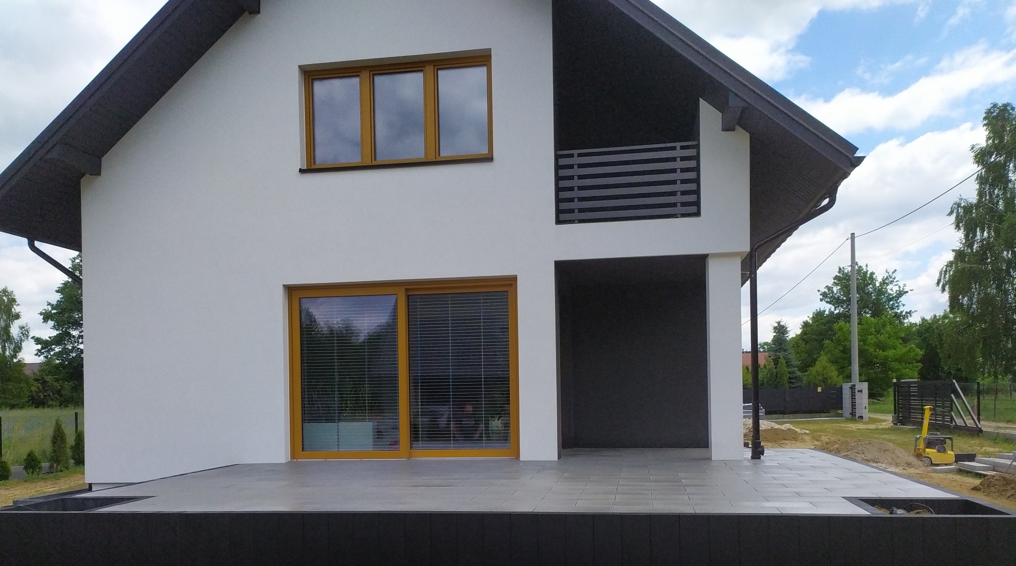 Nowoczesny dom z tarasem wyłożonym szarą kostką brukową. Widoczne detale wykończenia, jasna elewacja i elementy małej architektury. W tle zieleń drzew i błękitne niebo.