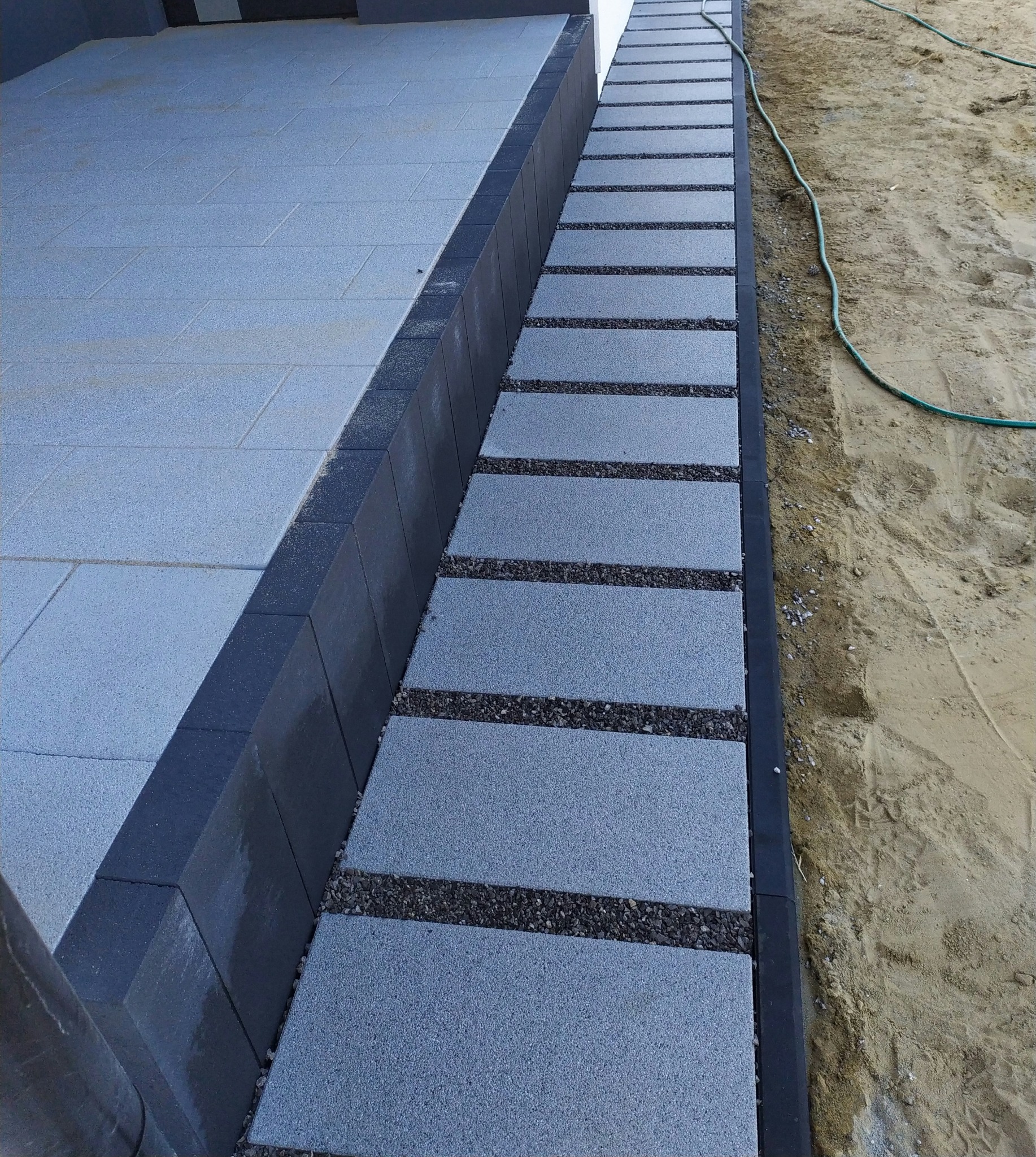 Szara kostka brukowa ułożona wzdłuż ścieżki z drobnym żwirem, obrzeża z ciemnej kostki. Widok z góry, w tle piasek i zielony wąż ogrodowy.