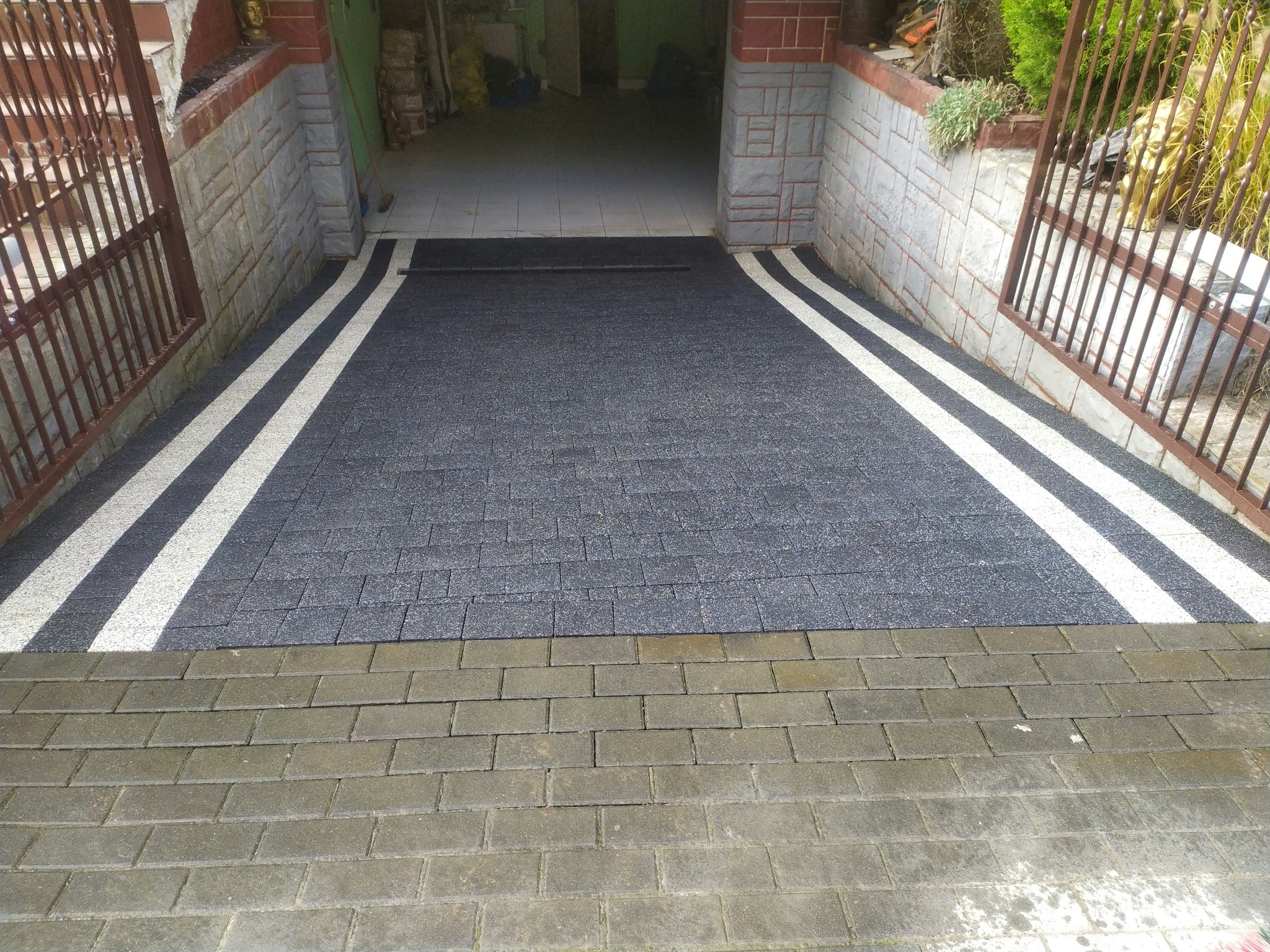 Nowa, ciemnoszara kostka brukowa z białymi pasami wjazdowymi, prowadząca do garażu. Widoczne bramy i fragment starszej kostki przed wjazdem.
