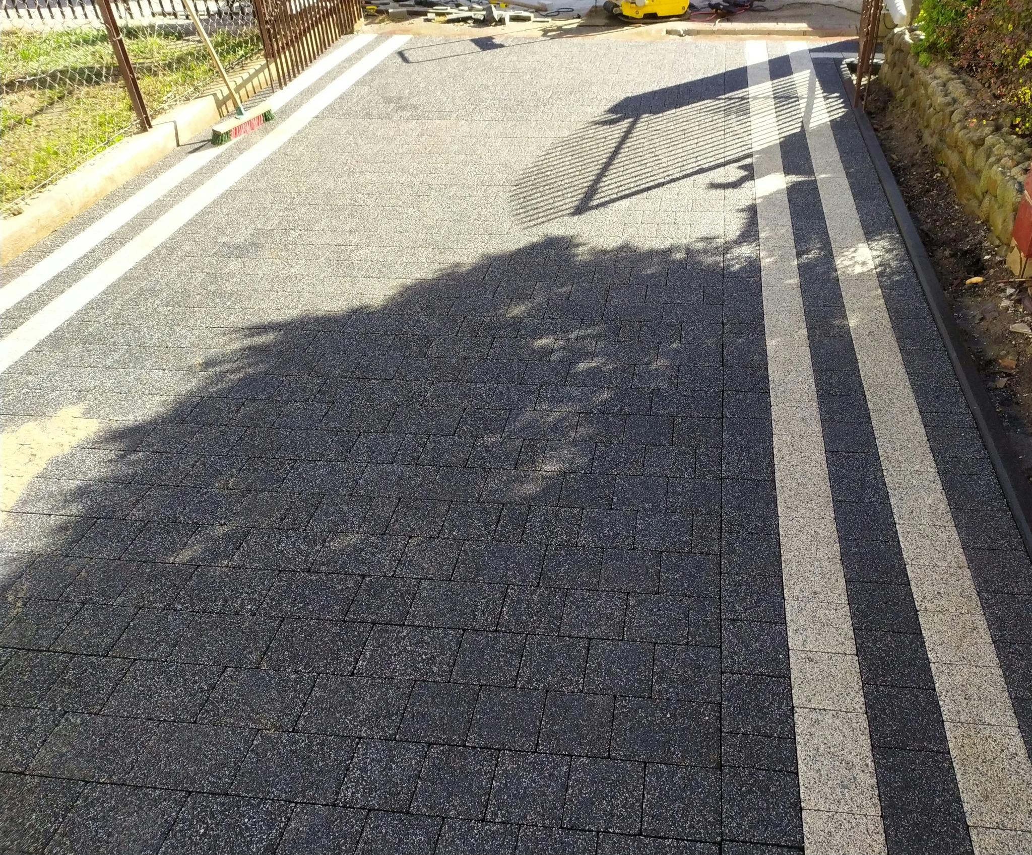 Ukończona nawierzchnia z kostki brukowej w odcieniach szarości, z jasnymi pasami. Widoczne cienie drzew i sprzęt budowlany w tle. Detal realizacji.