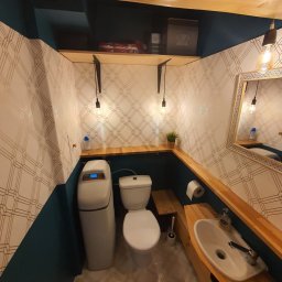 martin zych - Wnętrze wykończonej toalety z filtrem do wody, ściany w geometryczne wzory, drewniane półki i elementy wykończeniowe, oświetlenie w stylu retro.