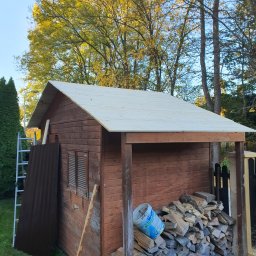 martin zych - Brązowy drewniany domek narzędziowy z nowym jasnym dachem z desek, oparty o ścianę blacha falista, obok sterta drewna i wiadro po farbie, na tle drzew.