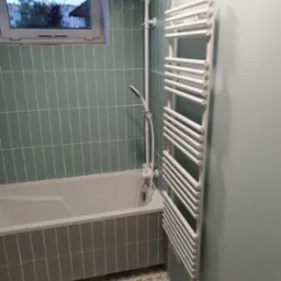 Nowoczesna łazienka z wanną obłożoną szarymi płytkami, zielonymi płytkami na ścianie, grzejnikiem drabinkowym i podłogą w kwiatowy wzór.