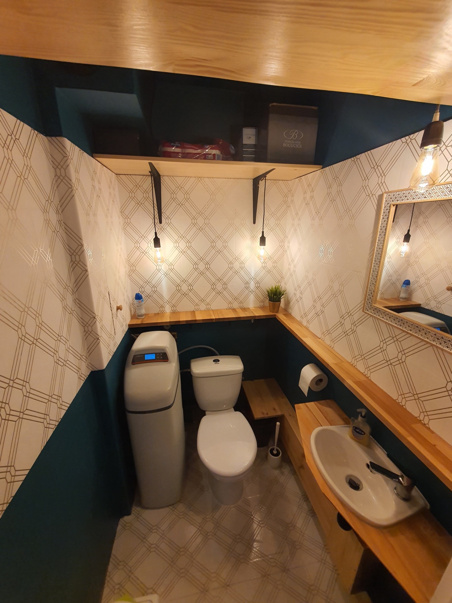 Wnętrze wykończonej toalety z filtrem do wody, ściany w geometryczne wzory, drewniane półki i elementy wykończeniowe, oświetlenie w stylu retro.