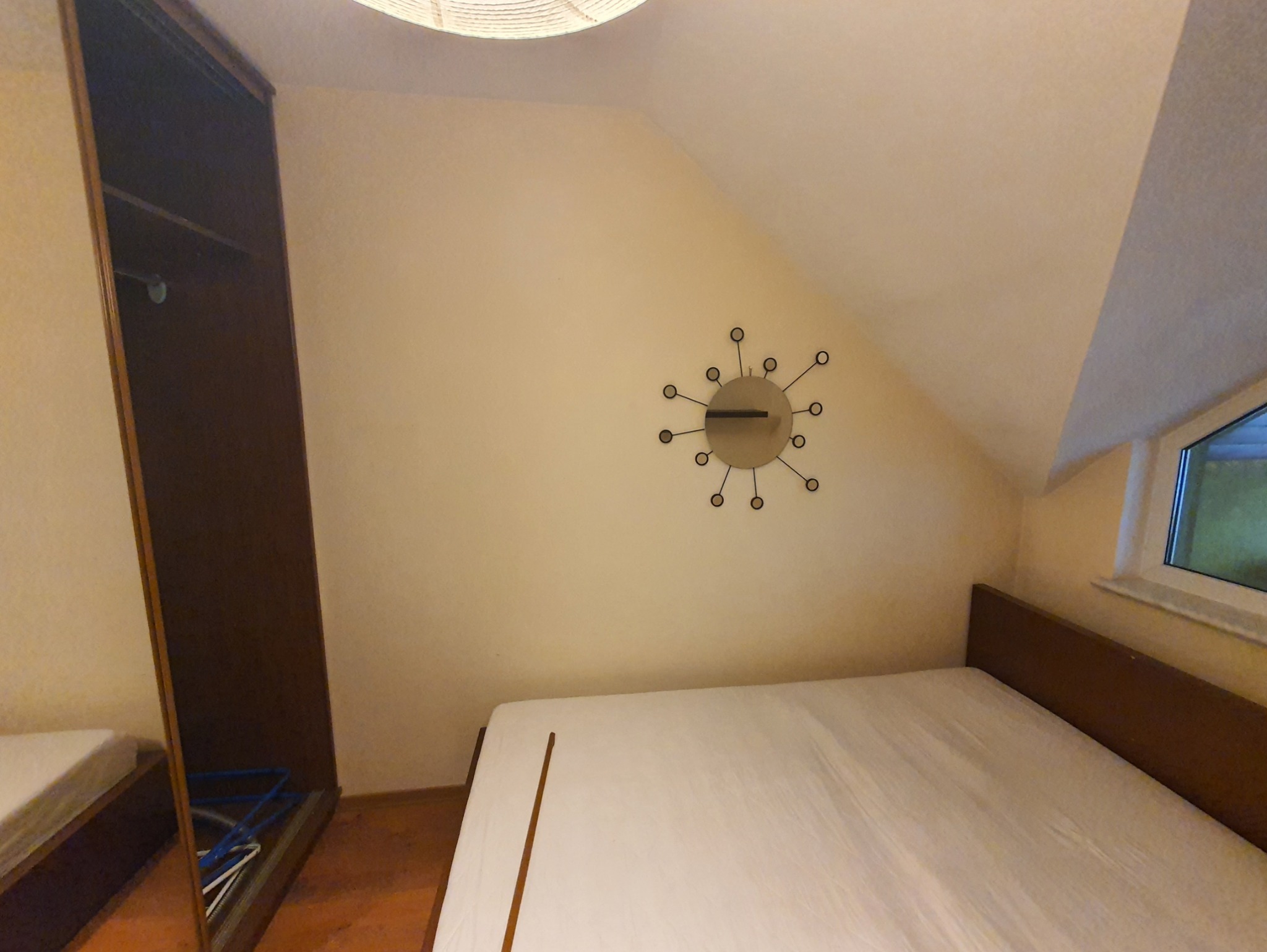 Pokój z łóżkiem z brązowym zagłówkiem, szafą z lustrem, zegarem w stylu retro na ścianie i oknem w skosie dachu.