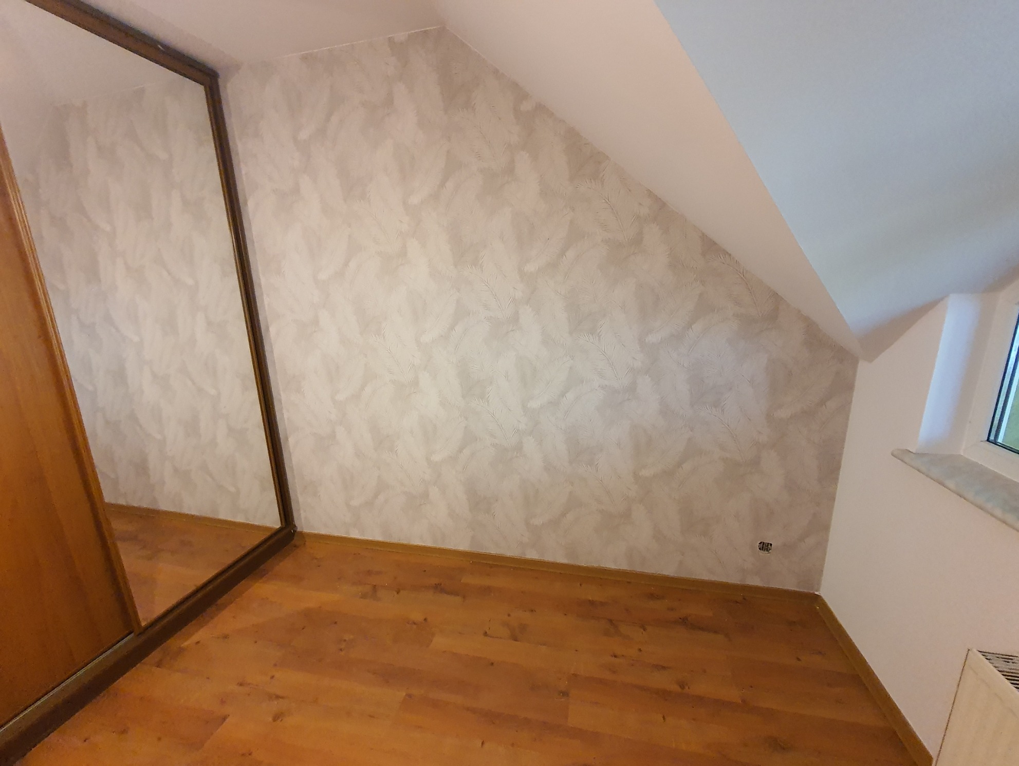 Pokój na poddaszu po remoncie: ściana w jasnych tapetach z motywem liści, drewniana podłoga z panelami, zabudowana szafa z lustrem i okno w skosie dachu.