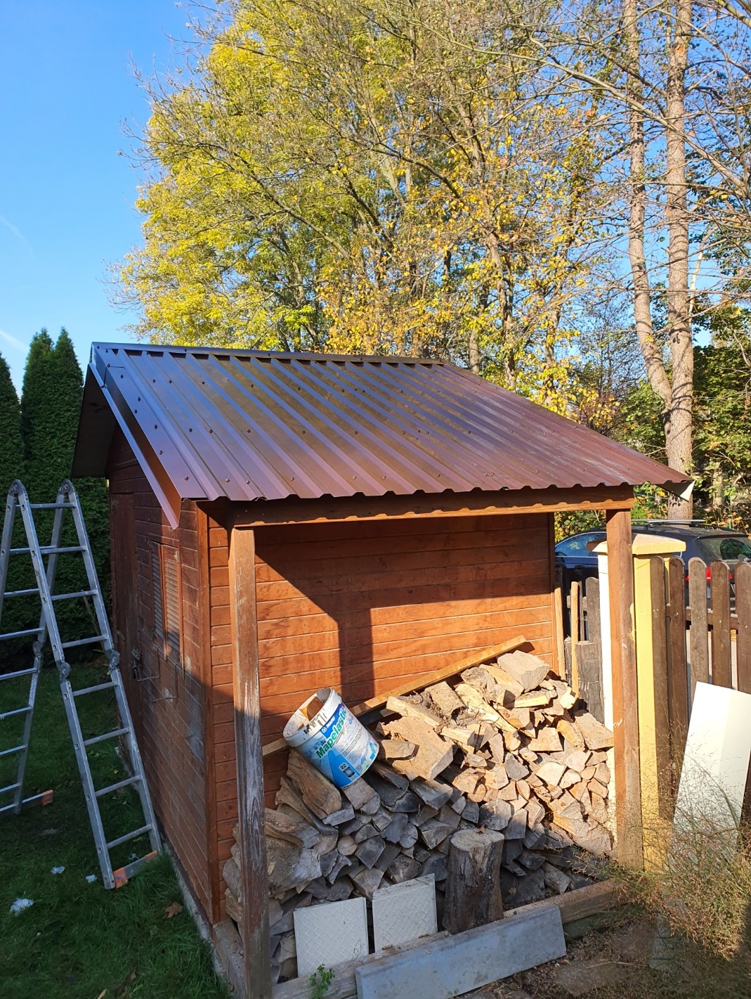 Brązowy blaszany dach na drewnianej szopie ogrodowej, z drabiną opartą o ścianę, stosem drewna i puszką po farbie obok, w słoneczny dzień.