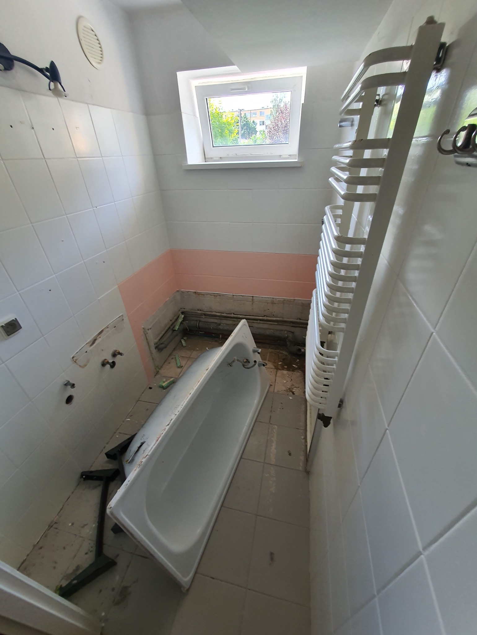 Demontaż wanny w łazience z częściowo usuniętymi płytkami, widoczne rury i grzejnik drabinkowy.