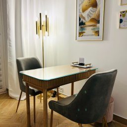 ORO Patrycja Andrzejczak-Solarz - Stylowe stanowisko do manicure z drewnianym biurkiem z szklanym blatem, dwoma szarymi krzesłami na złotych nogach i złotą lampą w tle, nad biurkiem wisi obraz z logo gabinetu kosmetycznego.