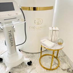 ORO Patrycja Andrzejczak-Solarz - Wnętrze gabinetu kosmetycznego: urządzenie Thesera Air In System, stolik z ampułkami Air-In i szklaną kulą dekoracyjną, na tle biała lada z logo Oro Beauty.