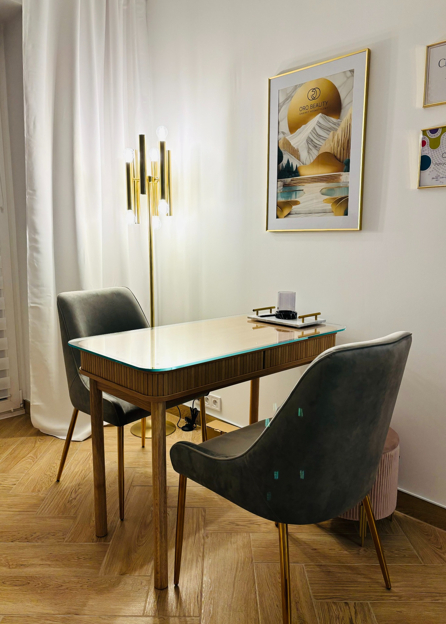 Stylowe stanowisko do manicure z drewnianym biurkiem z szklanym blatem, dwoma szarymi krzesłami na złotych nogach i złotą lampą w tle, nad biurkiem wisi obraz z logo gabinetu kosmetycznego.