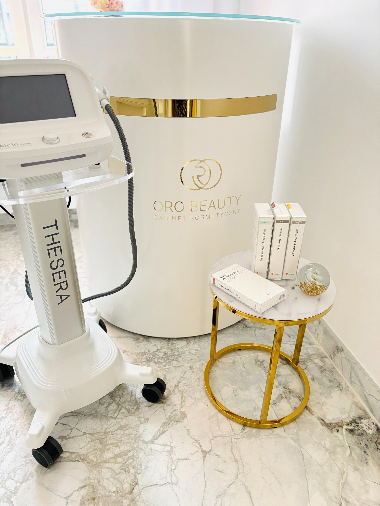 Wnętrze gabinetu kosmetycznego: urządzenie Thesera Air In System, stolik z ampułkami Air-In i szklaną kulą dekoracyjną, na tle biała lada z logo Oro Beauty.