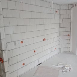 INSTALACJE ELEKTRYCZNE KRYSTIAN PIETRZAK - Surowa ściana z betonu komórkowego z widocznymi puszkami instalacyjnymi i przewodami elektrycznymi w trakcie montażu. Na podłodze leżą fragmenty kabli i karton.