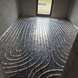 GAMA KLIMATYZAJE - Montaż ogrzewania podłogowego w surowym stanie: białe rurki ułożone w pętle na czarnej folii z siatką, widok z góry na nieotynkowane ściany i okno.