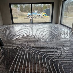 GAMA KLIMATYZAJE - Rozłożona instalacja ogrzewania podłogowego na folii izolacyjnej w surowym pomieszczeniu z widokiem na plac budowy za oknem.