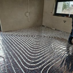 GAMA KLIMATYZAJE - Układanie rur grzewczych na folii aluminiowej z siatką w surowym wnętrzu domu jednorodzinnego, widoczna osoba w roboczym ubraniu.