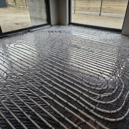 GAMA KLIMATYZAJE - Rozłożone rury ogrzewania podłogowego na folii izolacyjnej z siatką w surowym wnętrzu budynku z dużymi oknami.