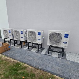 GAMA KLIMATYZAJE - Rząd jednostek zewnętrznych klimatyzacji Haier zamontowanych na czarnych wspornikach wzdłuż ściany budynku, z kartonem po urządzeniu leżącym na trawie.