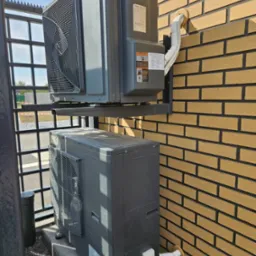 Dwa szare moduły urządzenia grzewczego zamontowane na zewnątrz budynku z cegły, jeden nad drugim, połączone rurami, widoczne elementy montażowe i otoczenie budynku.