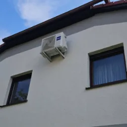 Biała jednostka zewnętrzna klimatyzatora marki Haier zamontowana na elewacji domu z dwoma oknami, widok z dołu na błękitne niebo.