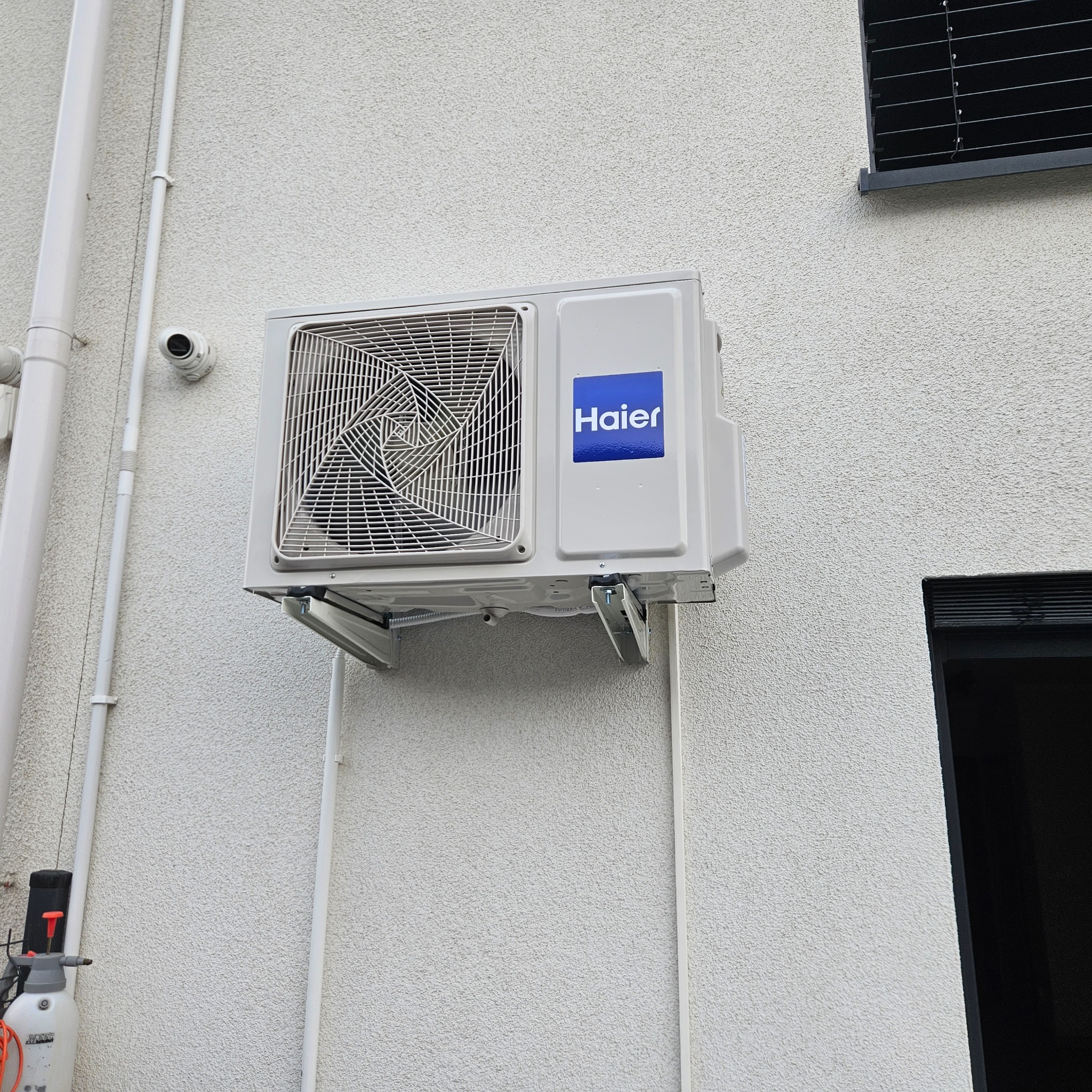 Biała zewnętrzna jednostka klimatyzacji marki Haier zamontowana na elewacji budynku z widocznymi przewodami i elementami instalacji.