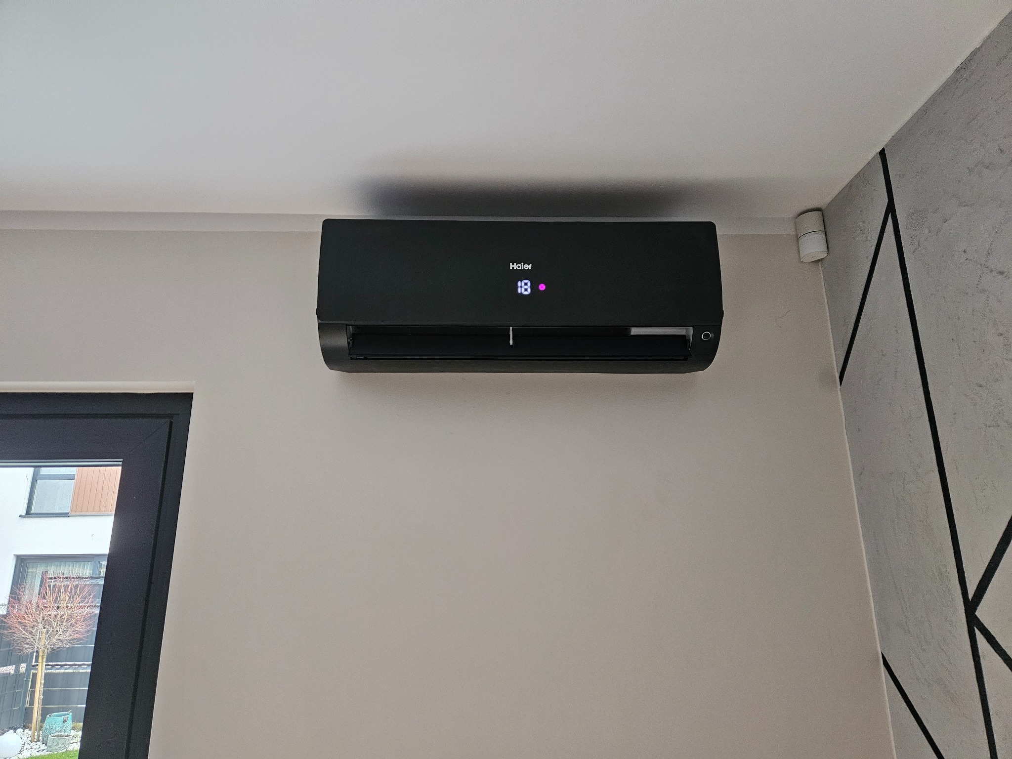 Czarny klimatyzator Haier zamontowany na ścianie w jasnym pomieszczeniu z widocznym oknem i fragmentem dekoracyjnej ściany.