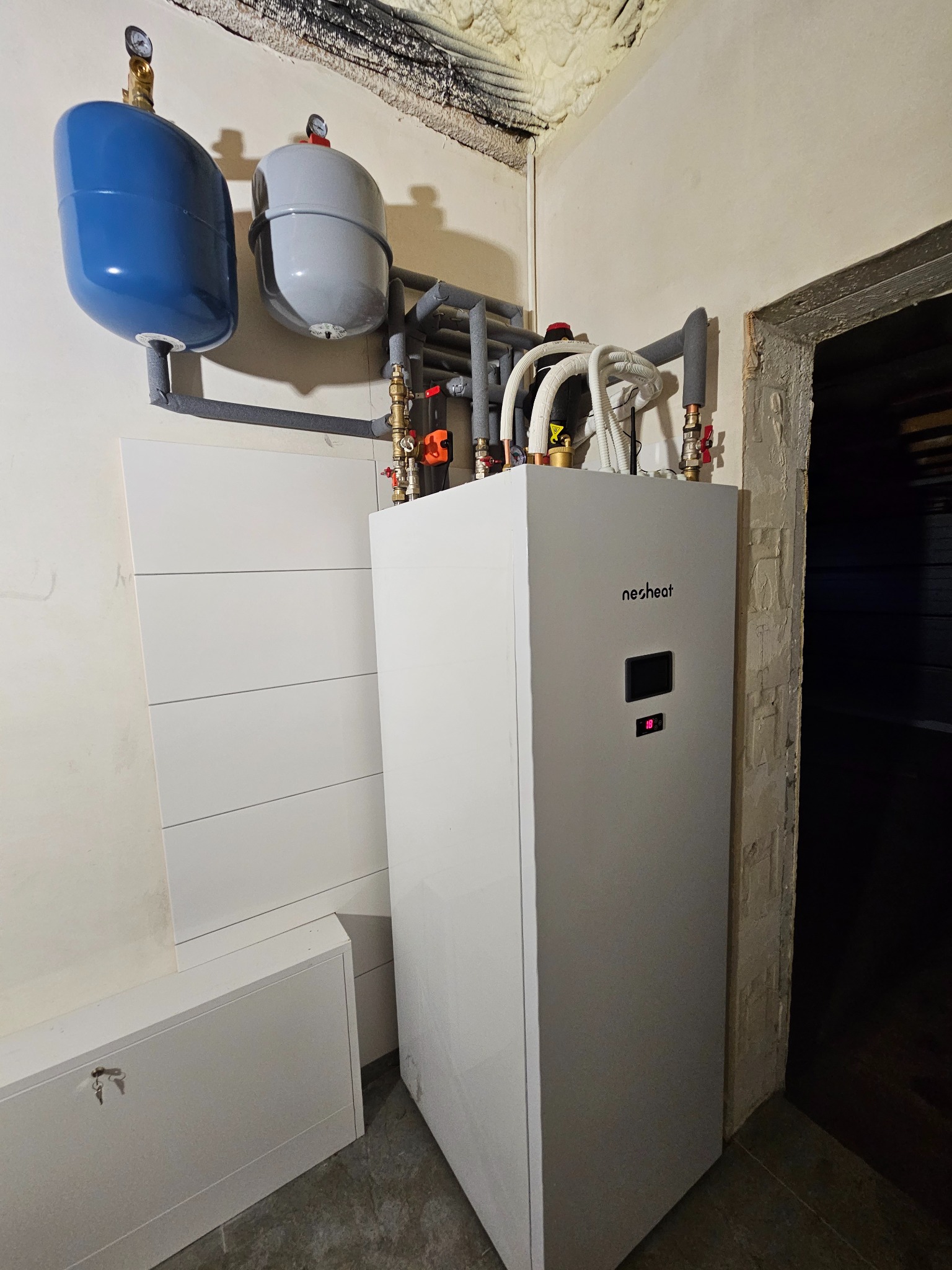 Biała pompa ciepła marki Nesheat z podłączonymi zbiornikami wyrównawczymi, rurami i zaworami, zamontowana przy ścianie z widoczną izolacją natryskową.