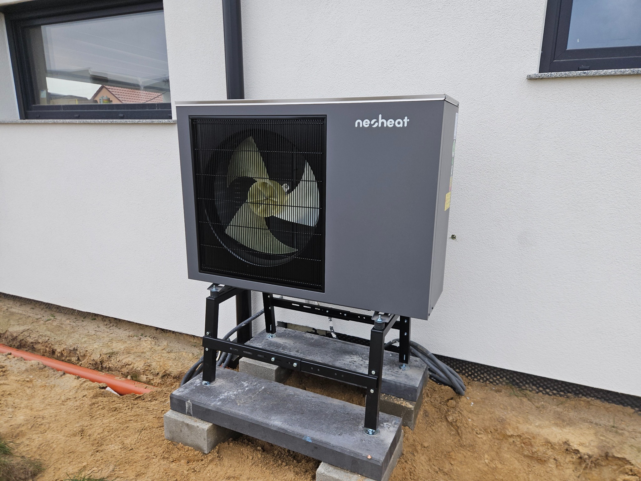 Szara pompa ciepła marki Nesheat zamontowana na metalowej konstrukcji z betonową podstawą przy białej ścianie budynku z widocznym oknem i pomarańczową rurą w ziemi.