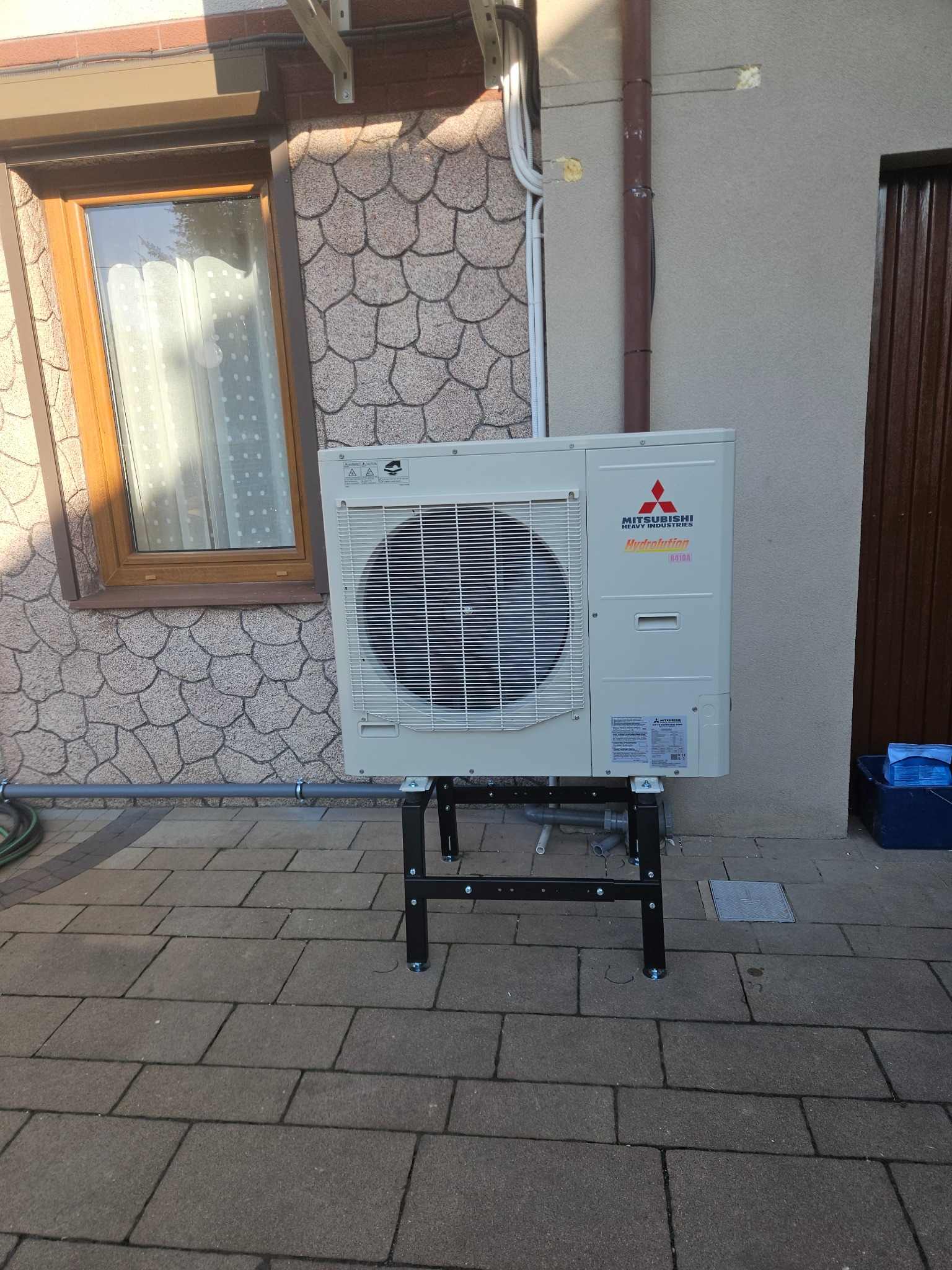 Zewnętrzna jednostka pompy ciepła Mitsubishi na czarnym metalowym stojaku, zainstalowana przy ścianie domu z oknem i dekoracyjną elewacją.