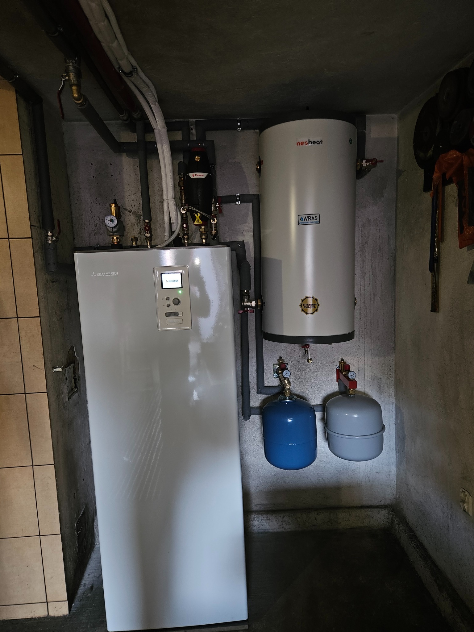 Biała pompa ciepła Mitsubishi, szary zbiornik Neosheat, dwa naczynia przeponowe (niebieskie i szare) oraz instalacja rurowa w kotłowni o surowych, betonowych ścianach.